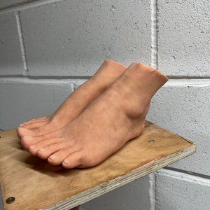 May include: Two realistic-looking flesh-toned feet made of silicone, resting on a wooden platform. The feet are positioned with the toes pointing towards the camera.
