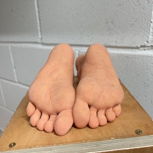 May include: A pair of realistic-looking silicone feet, with detailed toes and skin texture, are placed on a wooden platform.