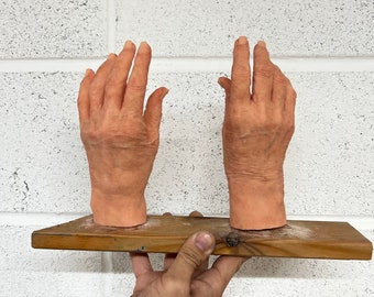 May include: Two realistic-looking, flesh-toned, silicone hands with detailed wrinkles and creases, resting on a wooden platform.