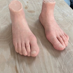 Pode incluir: Dois pés protéticos realistas feitos de um material de silicone cor de pele. Os pés estão posicionados sobre uma superfície de madeira clara.