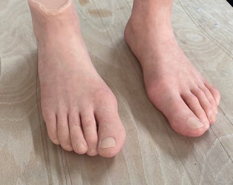 May include: Two realistic-looking prosthetic feet made of a flesh-toned silicone material. The feet are positioned on a light wood surface.