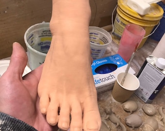 May include: A close-up of a realistic-looking silicone foot mold. The foot is being held by a hand and is resting on a pink mold. The mold is surrounded by other small, gray, clay-like objects.