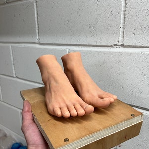 May include: Two realistic-looking, flesh-toned feet are mounted on a wooden platform. The platform is made of plywood and has a natural wood finish. The feet are positioned side-by-side, with the toes pointing forward.