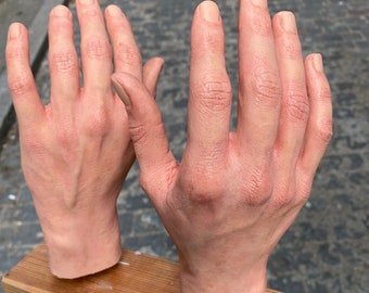 May include: Two realistic, flesh-toned hand sculptures are mounted on a wooden plank. The hands are detailed with visible veins and wrinkles, and the fingers are slightly curved. The wooden plank is light brown and appears to be made of multiple thin strips of wood.