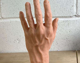 May include: A realistic-looking hand model made of silicone, with detailed veins and fingernails. The hand is posed with the palm facing up and fingers spread apart. The hand is resting on a wooden surface.