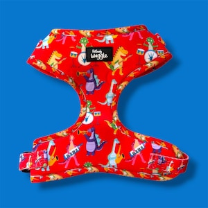 May include: A red pet harness featuring a repeating pattern of cartoon dinosaurs playing various musical instruments, including drums, guitars, and keyboards. A black rectangular label with the white text full body waggle is centered.
