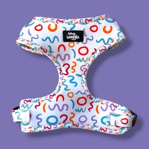 May include: A white pet harness featuring a colorful pattern of red, orange, blue, and purple squiggles and circles. A black rectangular label with the white text full body waggle is centered on the chest piece. The harness has black buckles.