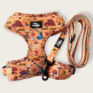 Puede incluir: Un arnés y correa para perros de color beige con un diseño de obra. El patrón incluye vehículos de construcción, trabajadores y conos de seguridad. El arnés tiene el texto "full body waggle". El conjunto está diseñado para combinar.