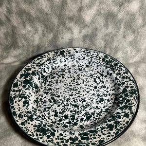 May include: A white enamel plate with a speckled green pattern and a black rim. The plate has a classic, rustic design, suitable for serving food. The speckled pattern adds a unique visual texture.