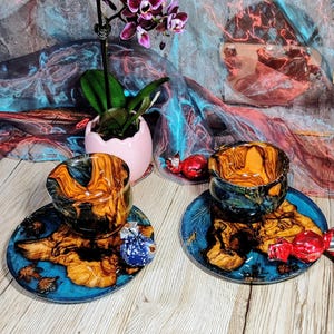 Può includere: Due set coordinati di tazze e piattini in legno fatti a mano. Le tazze e i piattini presentano una vibrante resina blu e un design a venature naturali del legno. Un piccolo vaso rosa con un'orchidea viola è sullo sfondo.