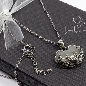 May include: A silver necklace with a floral pendant and a pale stone, presented on a dark grey gift box tied with a white ribbon. The text "Lovely Heart" is visible on the box.