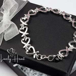 May include: A silver bracelet with heart and X-shaped links, resting on a black gift box with a white ribbon. The bracelet is a romantic jewelry piece. The box has the text "Lovely Heart".