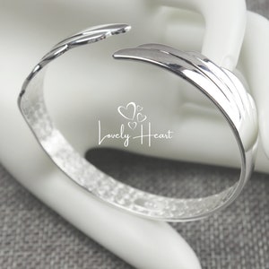 Może przedstawiać: Srebrna bransoletka typu bangle z fakturowanym wykończeniem i unikalnym wzorem w postaci zakrzywionego, warstwowego kształtu. Bransoletka jest otwarta i ma lekko młotkowany wygląd.