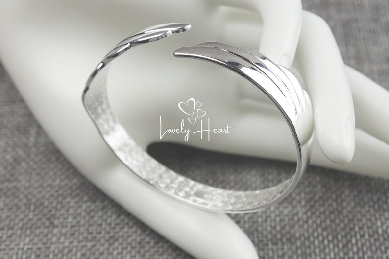 Może przedstawiać: Srebrna bransoletka typu bangle z fakturowanym wykończeniem i unikalnym wzorem w postaci zakrzywionego, warstwowego kształtu. Bransoletka jest otwarta i ma lekko młotkowany wygląd.