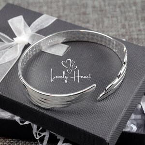 Może przedstawiać: Srebrna bransoletka typu bangle z wzorem skrzydeł. Bransoletka znajduje się w czarnym pudełku prezentowym z białym papierem jedwabnym.