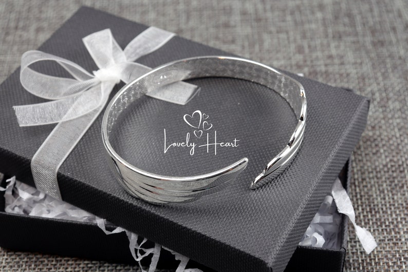 Może przedstawiać: Srebrna bransoletka typu bangle z wzorem skrzydeł. Bransoletka znajduje się w czarnym pudełku prezentowym z białym papierem jedwabnym.