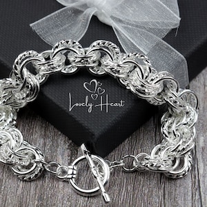 Peut inclure: Un bracelet en argent avec un fermoir à glissière. Le bracelet est composé de maillons épais et entrelacés. Le bracelet est posé sur une surface en bois.