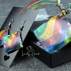May include: A rainbow-coloured necklace and earrings set. The necklace has a square pendant with a rainbow design and a multi-coloured cord. The earrings are also square with a rainbow design. The set is displayed on a black background with the text "Lovely Heart".