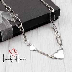 May include: A silver necklace featuring two heart pendants and a chain with oval links. The necklace is presented on a black gift box with a white ribbon.
