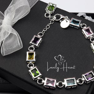 Könnte beinhalten: Ein silbernes Armband mit einer Kette und quadratischen Edelsteinen in verschiedenen Farben, darunter Grün, Pink, Blau und Gelb. Das Armband befindet sich in einer schwarzen Geschenkbox mit weißem Seidenpapier.