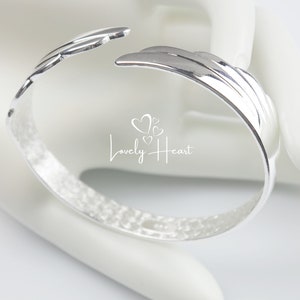 Może przedstawiać: Srebrna bransoletka z fakturowanym wykończeniem i wzorem skrzydła. Bransoletka jest otwarta i ma szczelinę po jednej stronie.