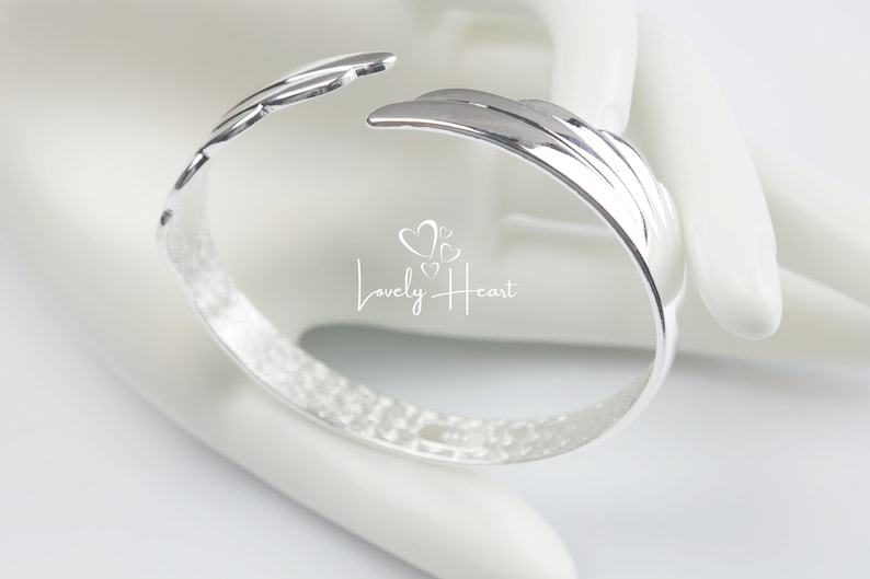 Może przedstawiać: Srebrna bransoletka z fakturowanym wykończeniem i wzorem skrzydła. Bransoletka jest otwarta i ma szczelinę po jednej stronie.