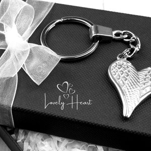 May include: A silver heart-shaped keychain with angel wings, presented in a black gift box with a white ribbon. The box has the text "Lovely Heart" printed on it.