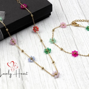 May include: A gold chain necklace and bracelet with colourful flower charms. The necklace is on a black box and the bracelet is on a wooden surface.