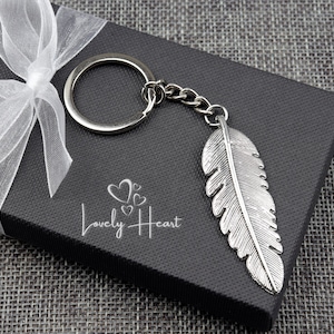 May include: Silver feather keyring with a silver ring and chain. The keyring is in a black gift box with a white ribbon and the words "Lovely Heart" printed on the box.