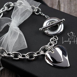 May include: A silver chain bracelet with a large heart charm. The bracelet has a toggle clasp and is displayed on a black box with the text "Lovely Heart".
