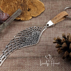 May include: Silver metal angel wing keyring with a cork and metal key ring. The keyring is on a brown hessian background.