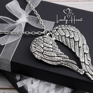 May include: A silver necklace with a pendant featuring two angel wings. The wings are detailed with intricate feather designs. The necklace is displayed in a black gift box with a white ribbon.