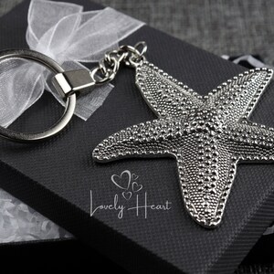 Op de afbeelding: Zilveren zeester-sleutelhanger met een zilveren ring en ketting. De sleutelhanger zit in een zwarte cadeaudoos met wit tissuepapier.