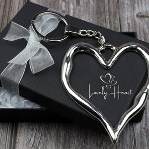 Op de afbeelding: Een zilveren hartvormige sleutelhanger met een kleiner hartontwerp en de tekst "Lovely Heart" erop gegraveerd. De sleutelhanger zit in een zwarte geschenkdoos met een wit lint.