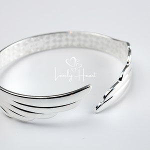 Może przedstawiać: Srebrna bransoletka z wzorem skrzydeł. Bransoletka jest otwarta i ma fakturowane wykończenie.