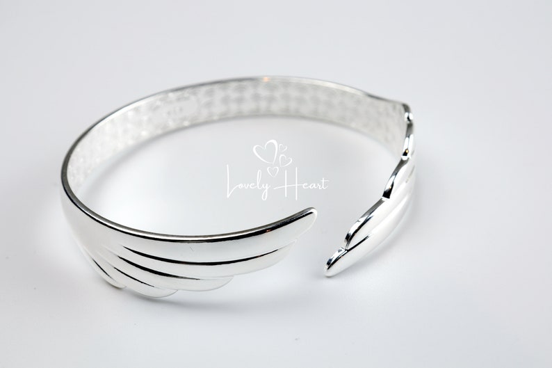 Może przedstawiać: Srebrna bransoletka z wzorem skrzydeł. Bransoletka jest otwarta i ma fakturowane wykończenie.