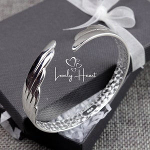 Może przedstawiać: Srebrna bransoletka typu bangle z fakturowanym wzorem. Bransoletka jest otwarta i ma zakrzywiony kształt. Bransoletka znajduje się w czarnym pudełku z białym papierem jedwabnym.