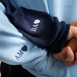 Op de afbeelding: Een lichtblauw sweatshirt met een donkerblauwe mouw. De mouw heeft het woord "BA" geborduurd met een klein wit hartje. De andere mouw heeft het woord "AM" geborduurd met een klein wit hartje.