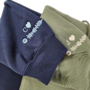 May include: Two hoodies, one navy blue and one olive green, with a heart and a music note followed by a sound wave graphic on each. The navy blue hoodie has a light blue heart and the olive green hoodie has a beige heart.