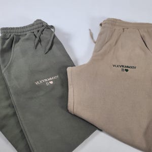 May include: Two pairs of sweatpants, one in olive green and one in beige, both with the same embroidered text "VLXV.VIII.MMXIV B" and a small heart.