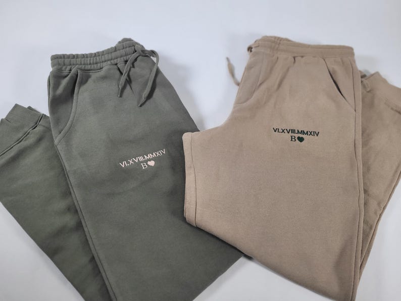 May include: Two pairs of sweatpants, one in olive green and one in beige, both with the same embroidered text "VLXV.VIII.MMXIV B" and a small heart.