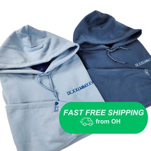 Op de afbeelding: Twee hoodies in lichtblauw en marineblauw. Elke hoodie heeft een trekkoord en het Romeinse cijfer "IX.XXI.MMXXV" geborduurd op de voorkant. De lichtblauwe hoodie heeft "ICT" en een hartsymbool op de mouw geborduurd.