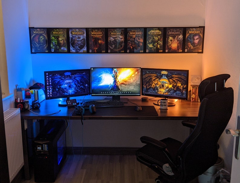 May include: A gaming setup with three monitors, a black gaming chair, and a computer tower. The monitors are displaying different images, including a dragon, a fantasy scene, and a dark background. The desk is made of wood and has a black mousepad. There are several World of Warcraft game boxes on the wall behind the monitors.