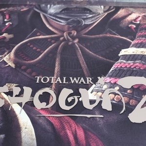 Puede incluir: Un primer plano de la portada de un videojuego que muestra a un guerrero samur&aacute;i con armadura tradicional. El t&iacute;tulo "TOTAL WAR: SHOGUN 2" est&aacute; escrito en una fuente estilizada.