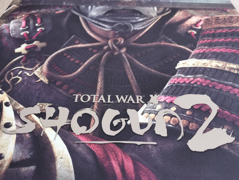 Puede incluir: Un primer plano de la portada de un videojuego que muestra a un guerrero samur&aacute;i con armadura tradicional. El t&iacute;tulo "TOTAL WAR: SHOGUN 2" est&aacute; escrito en una fuente estilizada.