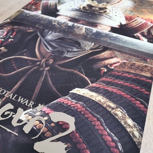 Puede incluir: Un p&oacute;ster para el videojuego Total War: Shogun 2. El p&oacute;ster presenta a un guerrero samur&aacute;i con armadura completa, sosteniendo una espada katana. El rostro del guerrero est&aacute; oculto por la espada, pero su armadura es visible, incluyendo un casco rojo y dorado y una coraza negra y dorada. El p&oacute;ster est&aacute; situado sobre un fondo de campo de batalla, con humo y fuego en el fondo.