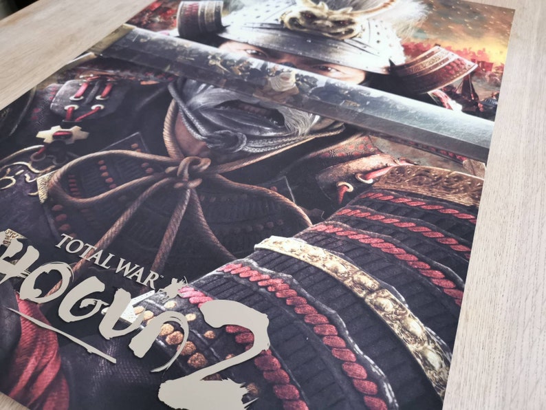 Puede incluir: Un p&oacute;ster para el videojuego Total War: Shogun 2. El p&oacute;ster presenta a un guerrero samur&aacute;i con armadura completa, sosteniendo una espada katana. El rostro del guerrero est&aacute; oculto por la espada, pero su armadura es visible, incluyendo un casco rojo y dorado y una coraza negra y dorada. El p&oacute;ster est&aacute; situado sobre un fondo de campo de batalla, con humo y fuego en el fondo.