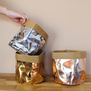 May include: Three metallic paper storage bags in silver, gold, and rose gold. Each bag has a brown paperboard rim. One bag is being held by a hand. The bags are stacked on top of each other.
