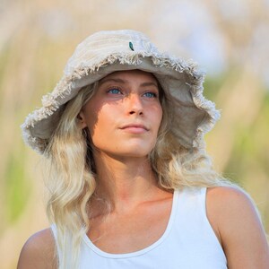 May include: A person wearing a beige sun hat with a frayed edge and a small green detail. They are also wearing a white vest top and dark blue athletic trousers. The background is blurred with natural colours.