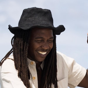 May include: A black hat with a wide brim is worn by a person with long dreadlocks. The person is wearing a light-coloured button-down shirt. The background is a bright blue sky.